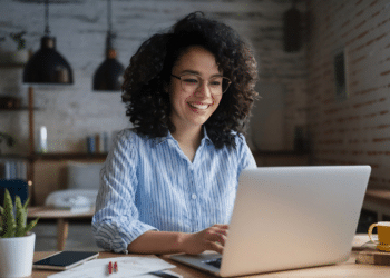 Mulher de óculos usando notebook e sorrindo em ambiente doméstico iluminado