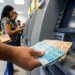 Pessoa segurando notas de R$ 100 e R$ 50 ao retirar dinheiro em caixa eletrônico no banco