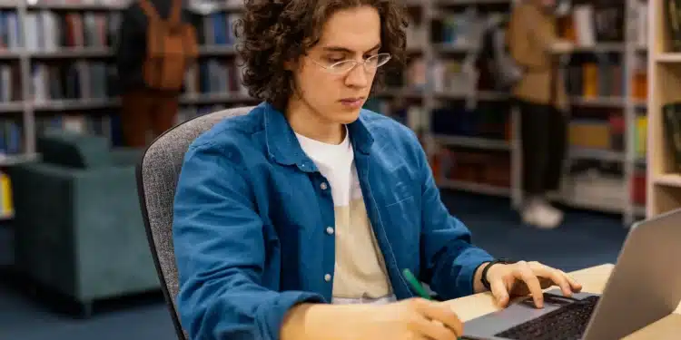 Estudante com laptop e caderno, focado no TCC em uma biblioteca.