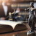 Estátua da Justiça com balança sobre mesa ao lado de livro aberto em biblioteca jurídica