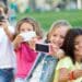 Quatro crianças tirando selfies com smartphones em parque, sorrindo e próximas umas das outras.