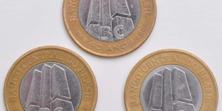 Fotografia de três moedas de 1 Real comemorativas dos 40 anos do Banco Central, organizadas em formato de triângulo sobre um fundo branco. A imagem exibe o lado do anverso das moedas, destacando os detalhes do núcleo prateado e as legendas douradas.