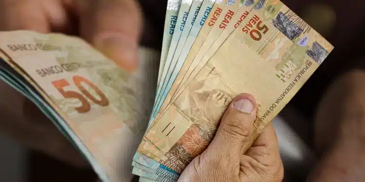 Mãos de idoso segurando e contando cédulas de reais em ambiente fechado.