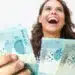 Mulher sorrindo segurando cédulas de 100 reais após anúncio de pagamento de R$ 950 pela Caixa para CPFs final 0 a 9