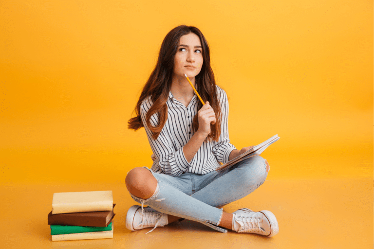 Jovem sentada no chão segura lápis e caderno, olha para cima pensativa ao lado de livros empilhados.