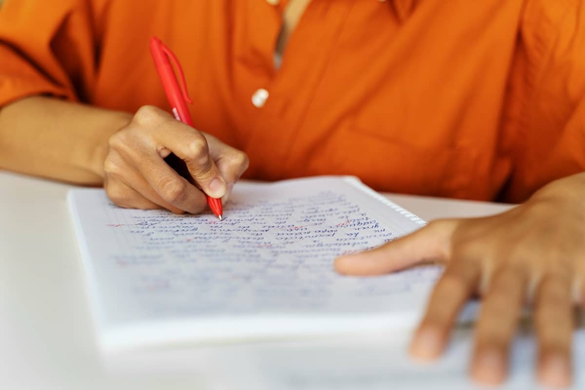 Pessoa de camisa laranja escrevendo em caderno com caneta vermelha sobre mesa branca