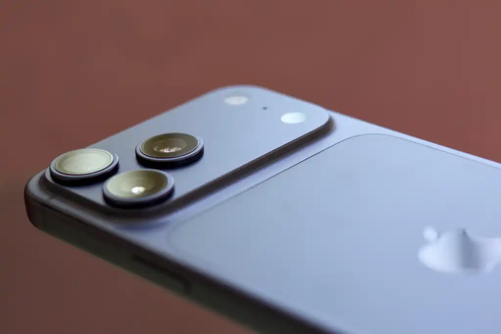 Close macro focado no módulo de três lentes traseiras e flash de um smartphone na cor azul-lavanda suave, simulando o design especulativo do iPhone 18 Pro. A imagem mostra detalhes do relevo das lentes sobre um fundo marrom desfocado.