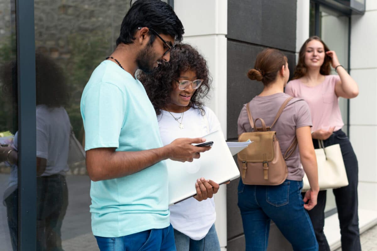 Dois jovens universitários consultando smartphone com notebook em frente a edifício moderno e outras duas estudantes ao fundo em área externa