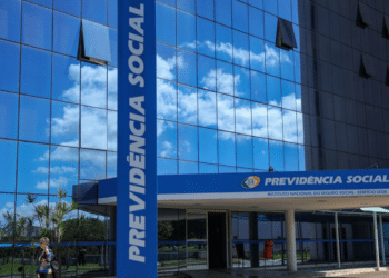 Entrada do prédio do INSS com letreiro azul da Previdência Social e reflexo de nuvens no vidro