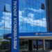 Entrada do prédio do INSS com letreiro azul da Previdência Social e reflexo de nuvens no vidro