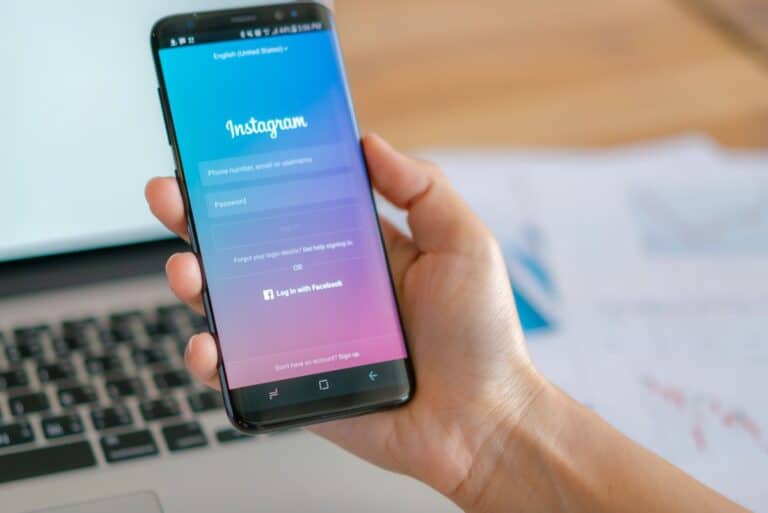 Mão segurando um smartphone com a tela de login do Instagram aberta sobre uma mesa de escritório com um notebook.