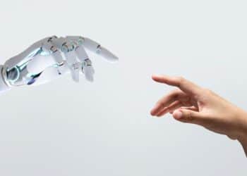 Close em plano médio de uma mão robótica branca e articulada e uma mão humana de pele morena se aproximando, quase se tocando, em um fundo cinza claro neutro. A imagem simboliza a interação entre humanos e inteligência artificial no mercado de trabalho.