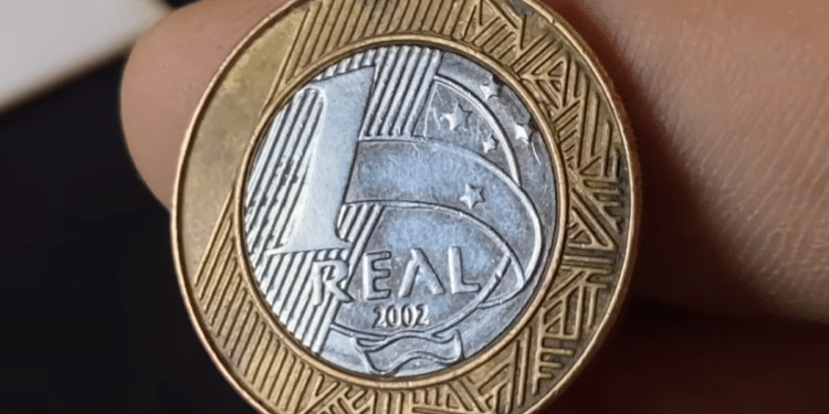 Foto em primeiro plano de uma moeda de 1 real brasileira datada de 2002, exibindo o reverso com o valor e o ano, segurada por dedos em um fundo escuro.