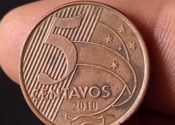 Close-up detalhado do reverso de uma moeda de 5 centavos de real do ano de 2010, sendo segurada pelos dedos de uma pessoa contra um fundo escuro.