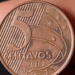 Close-up detalhado do reverso de uma moeda de 5 centavos de real do ano de 2010, sendo segurada pelos dedos de uma pessoa contra um fundo escuro.