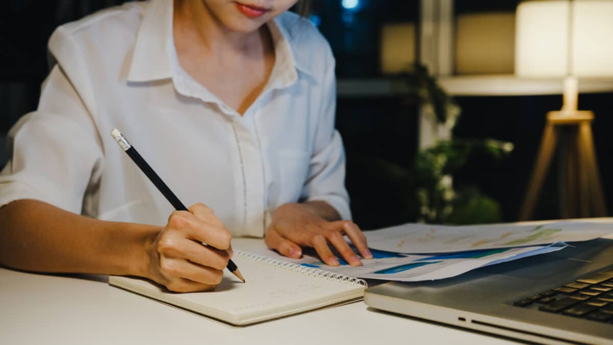 Jovem escrevendo em caderno à noite com documentos e notebook representando como estruturar a redação do Enem 2026