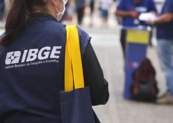 Mulher com colete do IBGE segura bolsa amarela enquanto aplica pesquisa em área urbana