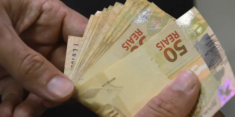 Mãos segurando diversas cédulas de cinquenta reais abertas em leque sobre fundo escuro