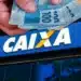 Pessoa segurando notas de R$ 100 em frente à agência da Caixa Econômica Federal, relacionada ao pagamento de R$ 800 para CPFs com finais de 0 a 9.