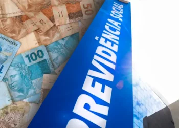Cédulas de 50 e 100 reais sobrepostas a placa azul com texto "Previdência Social" e prédio espelhado ao fundo.