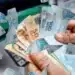 Mãos segurando cédulas de 100, 50 e 20 reais em frente a caixa eletrônico com notas voando