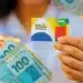 Cartão do Bolsa Família sendo segurado por uma pessoa, com cédulas de dinheiro de R$ 100, R$ 50 e R$ 20 ao fundo, representando os pagamentos do programa em abril de 2026.