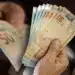 Pessoa segurando cédulas de real representando pagamentos de benefícios sociais em maio.