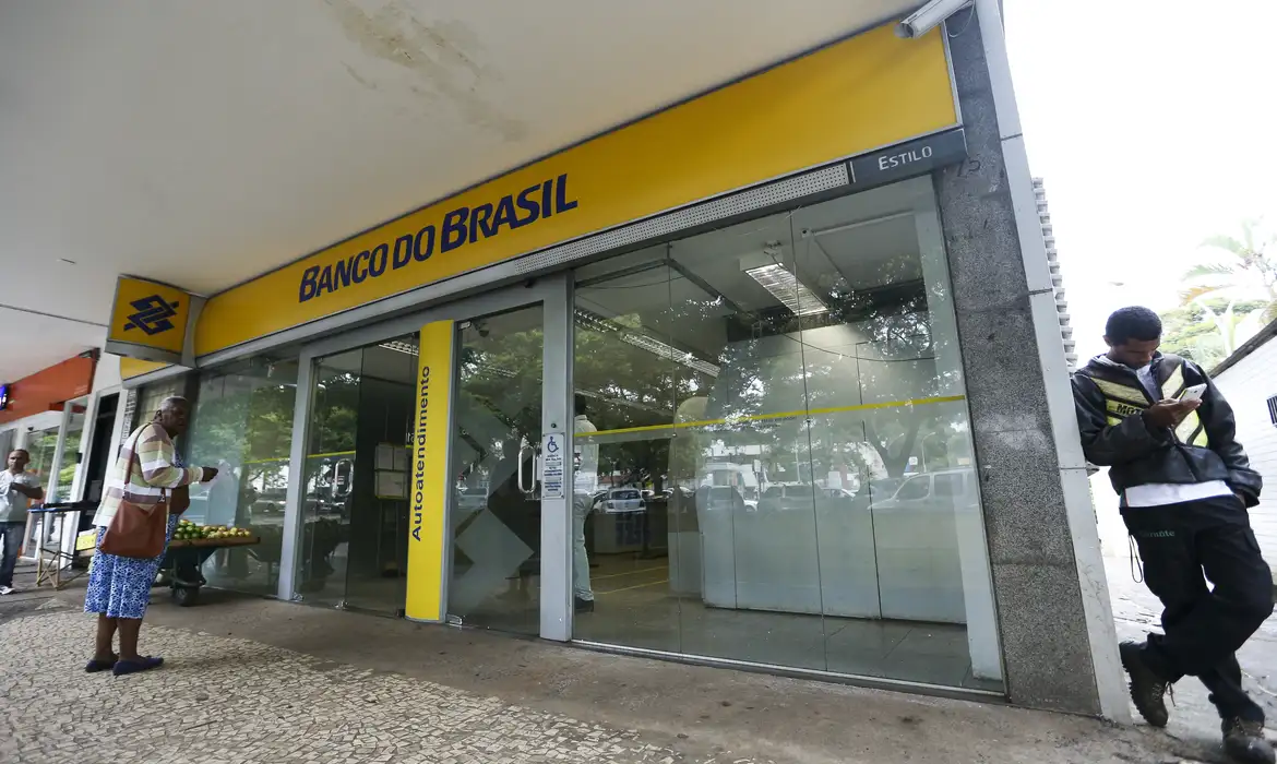 Agência Banco do Brasil com letreiro amarelo, portas de vidro, área de autoatendimento e pedestres na calçada