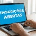 Mãos digitando em notebook prateado com tela azul exibindo "Inscrições Abertas", sobre mesa de madeira com caderno e caneca