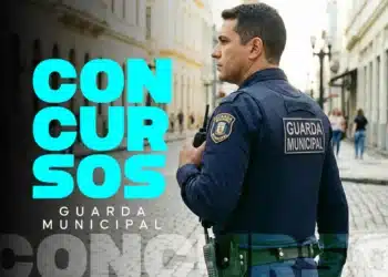 Guarda Municipal de uniforme azul-marinho segura rádio comunicador em rua de paralelepípedos com prédios coloniais ao fundo.