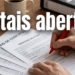 Mão segurando caneta vermelha sobre documentos de editais de concurso e processo seletivo em mesa de madeira