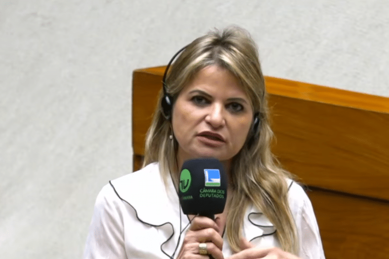 Mulher loira de cabelos longos ondulados, blusa branca com detalhes pretos, segura microfone da TV Câmara e Câmara dos Deputados