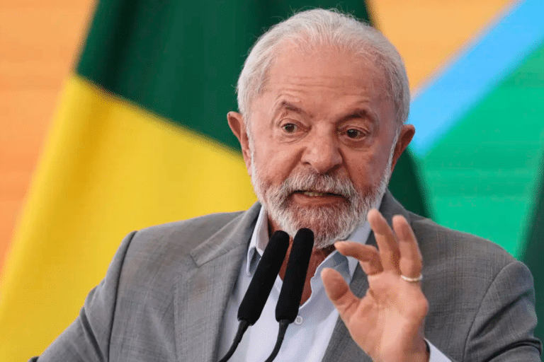Presidente Lula gesticula durante pronunciamento com terno cinza, camisa azul e bandeiras do Brasil ao fundo
