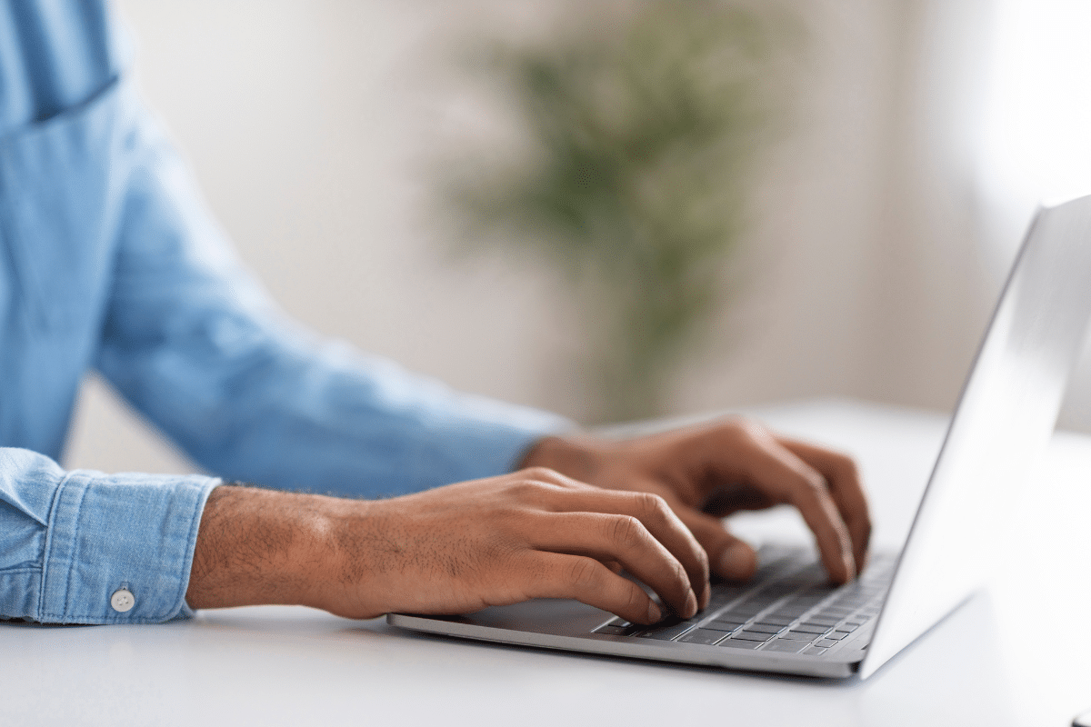 Mãos de homem com camisa jeans azul digitando no teclado de notebook prata em mesa branca com planta ao fundo