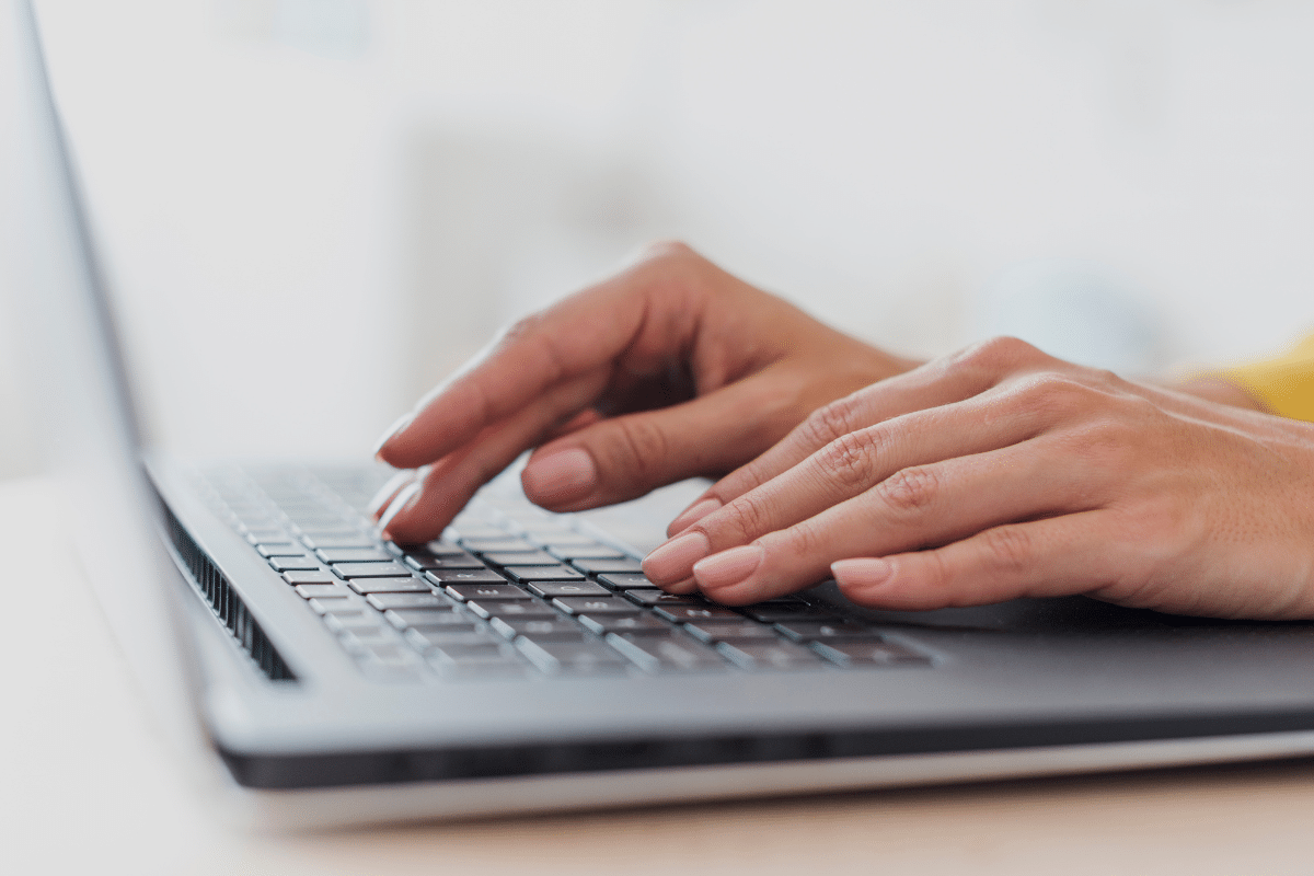 Mãos com unhas naturais digitando em teclado de laptop cinza sobre mesa branca
