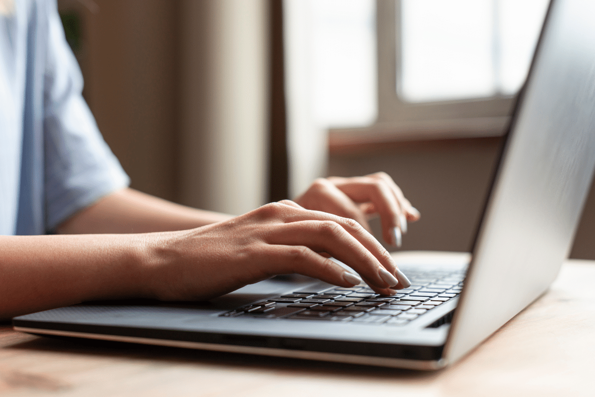 Mãos femininas com unhas claras digitando em teclado de notebook prateado sobre mesa de madeira clara