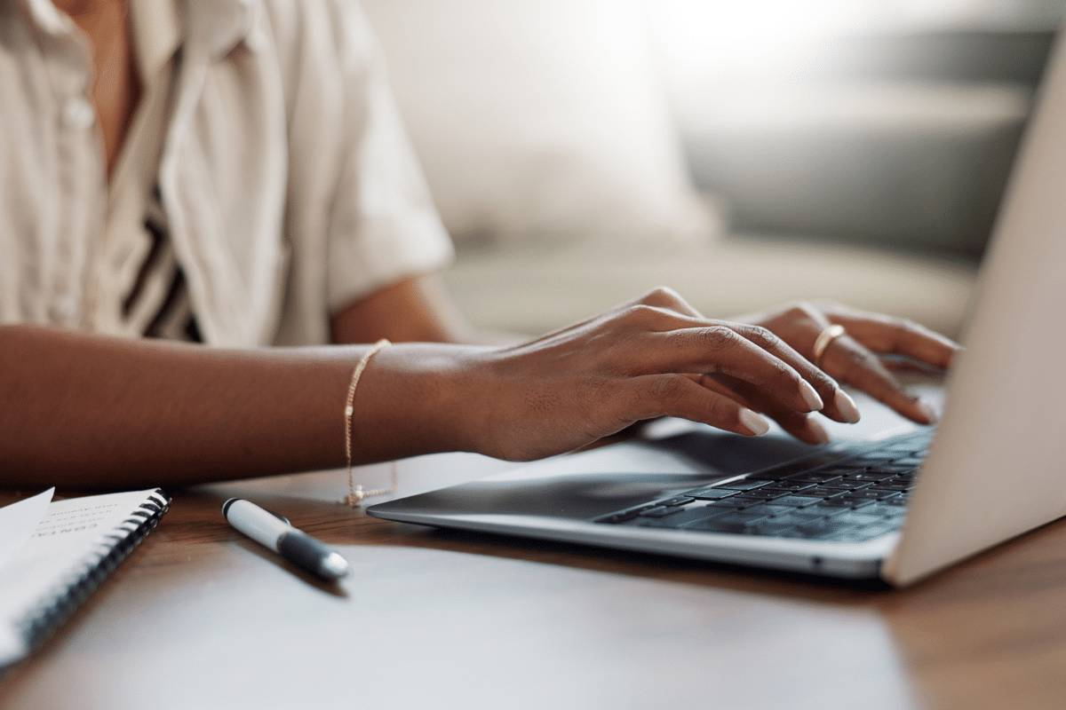Mãos de mulher negra com anel dourado e pulseira digitando em notebook prata sobre mesa de madeira com caderno
