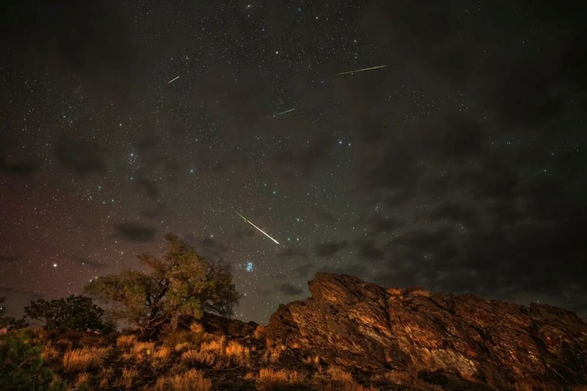Chuva de meteoros Líridas iluminando o céu estrelado durante a madrugada em área de natureza com rochas e árvores.