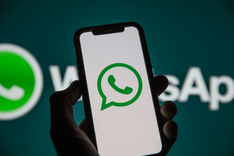 Mão segura celular preto mostrando ícone verde do WhatsApp, com logo ampliado ao fundo