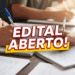 Mãos de estudantes escrevendo em cadernos sobre mesa de madeira com destaque vermelho "Edital Aberto!"