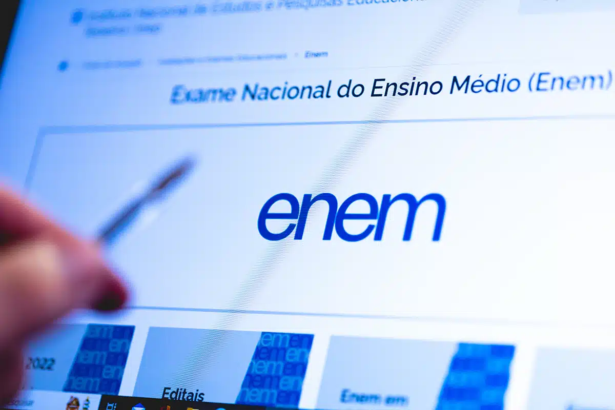 Tela do site do Enem exibida em computador com pessoa utilizando caneta, representando preparação e pesquisa sobre atualidades para o exame.