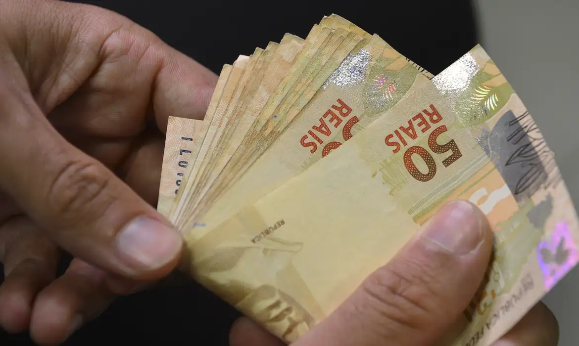 Mãos segurando leque de notas de R$ 50 representando o reajuste do salário mínimo 2027