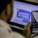 Homem consulta no celular o aplicativo da Receita Federal enquanto a página do Imposto de Renda 2026 está aberta no notebook ao fundo.
