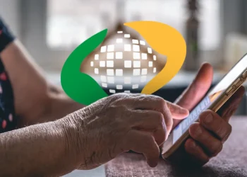 Mãos enrugadas de idosa segurando smartphone dourado em frente ao logotipo do INSS