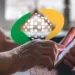 Mãos enrugadas de idosa segurando smartphone dourado em frente ao logotipo do INSS