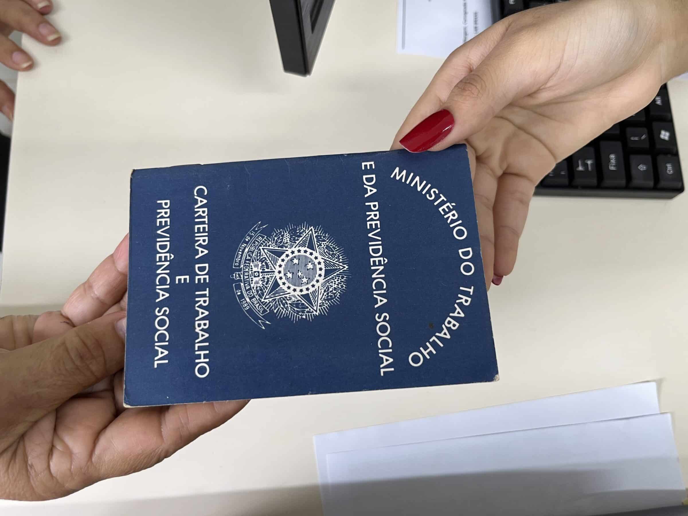 Mãos femininas seguram CTPS azul do Ministério do Trabalho com brasão dourado sobre mesa de escritório.