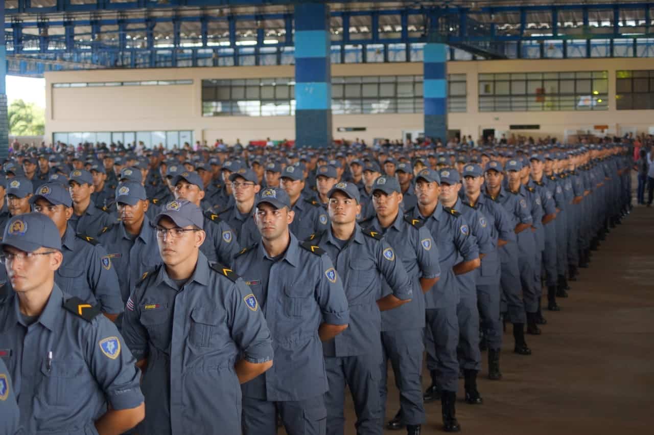 Centenas de soldados da PMMA em formação, uniformes cinza-azulados, bonés e brasões, em ginásio coberto.