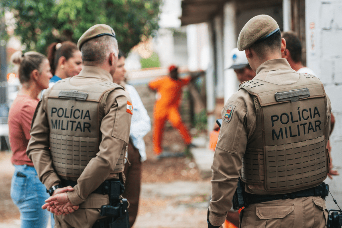 Dois policiais militares de costas com coletes bege observam civis e trabalhador de laranja em rua