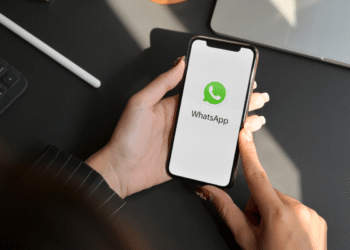 Mãos femininas com unhas vermelhas segurando iPhone exibindo logo do WhatsApp em ambiente de escritório