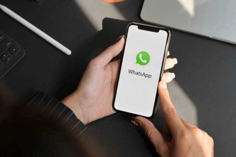 Mãos femininas com unhas vermelhas segurando iPhone exibindo logo do WhatsApp em ambiente de escritório